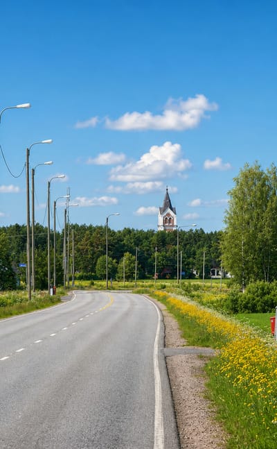 Road leading into Lumijoki village with the church tower rising in the distance among summer greenery.