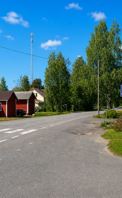 A quiet rural crossroads with a small red cottage and white timber house beside a telecom mast in Merijärvi, Finland.
