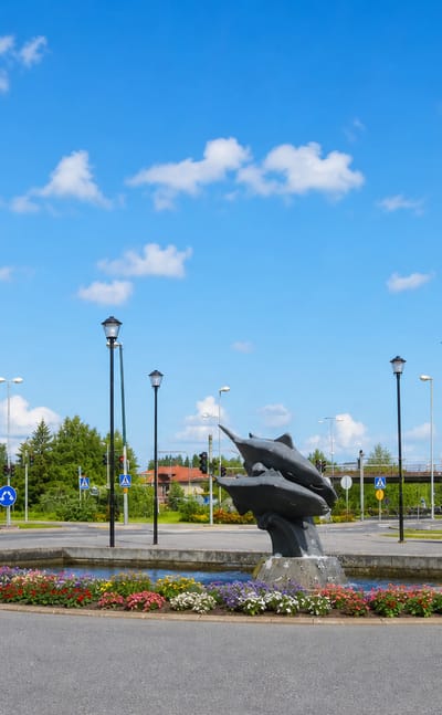 Roundabout in Muhos, Finland with a leaping-fish sculpture, flower beds and a tall spruce tree.