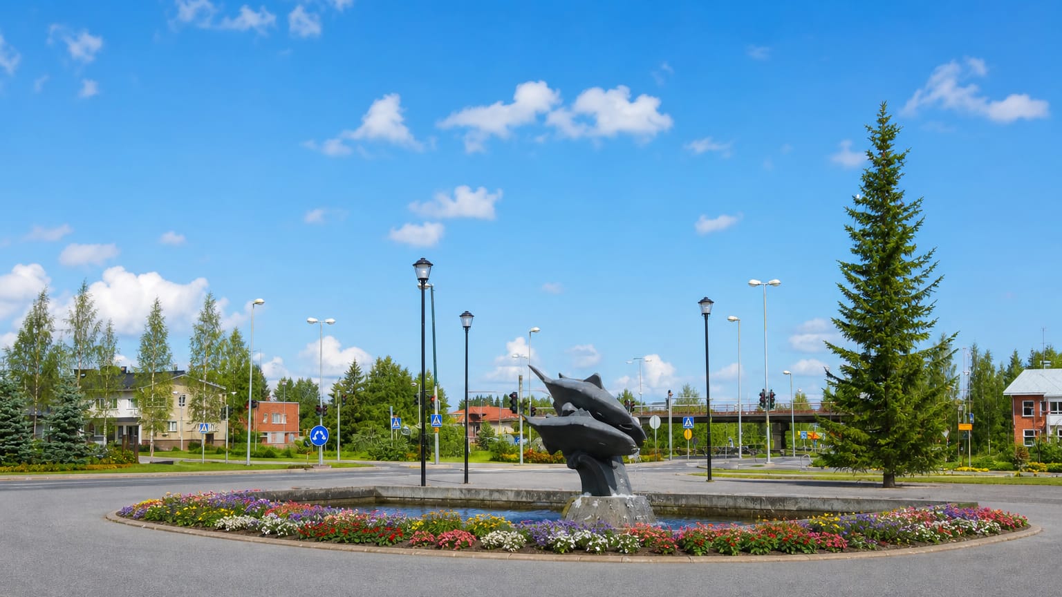 Roundabout in Muhos, Finland with a leaping-fish sculpture, flower beds and a tall spruce tree.