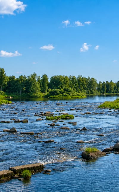 Rocky river rapids through Oulainen, Finland, with forested banks and a red riverside building under a blue summer sky.