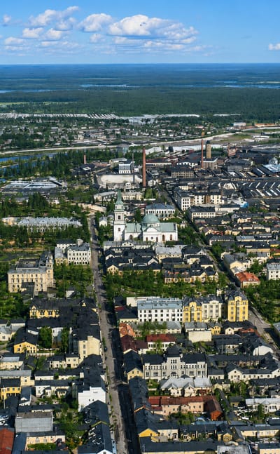 Aerial daytime view of Oulu, Finland: riverside grid of low buildings and a church spire, with forests and sea beyond.