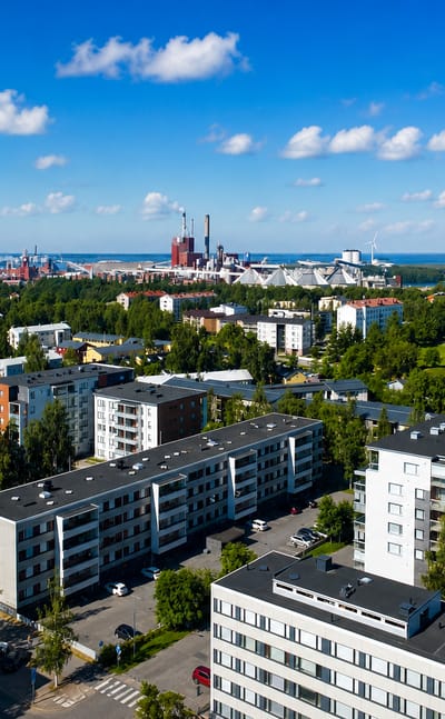 Aerial daytime view over Heinäpää in Oulu, Finland, with apartment blocks and seaside industrial chimneys on the horizon.