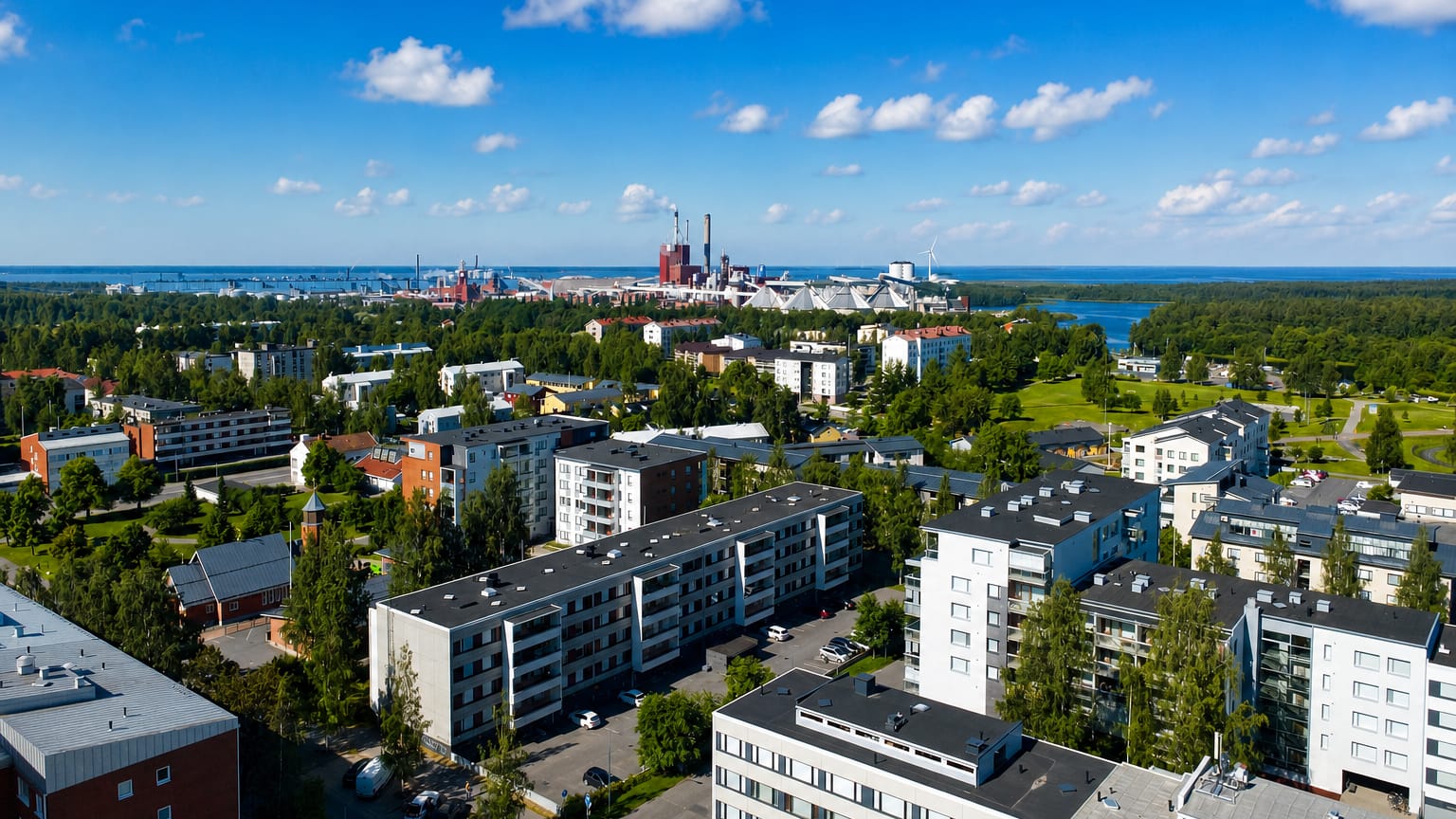 Aerial daytime view over Heinäpää in Oulu, Finland, with apartment blocks and seaside industrial chimneys on the horizon.