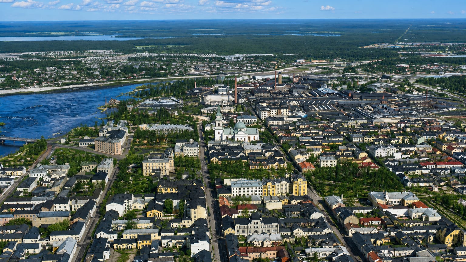 Aerial daytime view of Oulu, Finland: riverside grid of low buildings and a church spire, with forests and sea beyond.