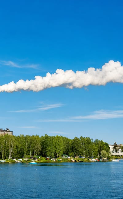 Tall power-plant chimney trailing a steam plume across the bay from the forested shores of Hietasaari, Oulu.
