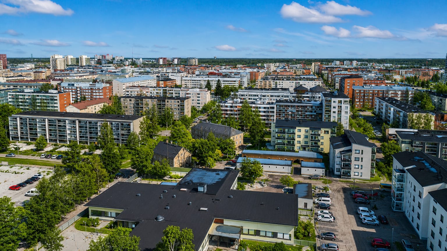 Aerial daytime view of the Hollihaka district in Oulu, Finland, with mid-rise apartment blocks, parking lots and leafy trees.
