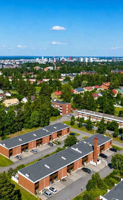 Aerial view of red-brick terraced housing and pine woods in Höyhtyä, with the Oulu skyline beyond.