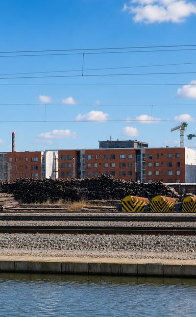 Red-brick apartment blocks behind electrified railway tracks, timber stacks and a canal in Limingantulli, Oulu, Finland.