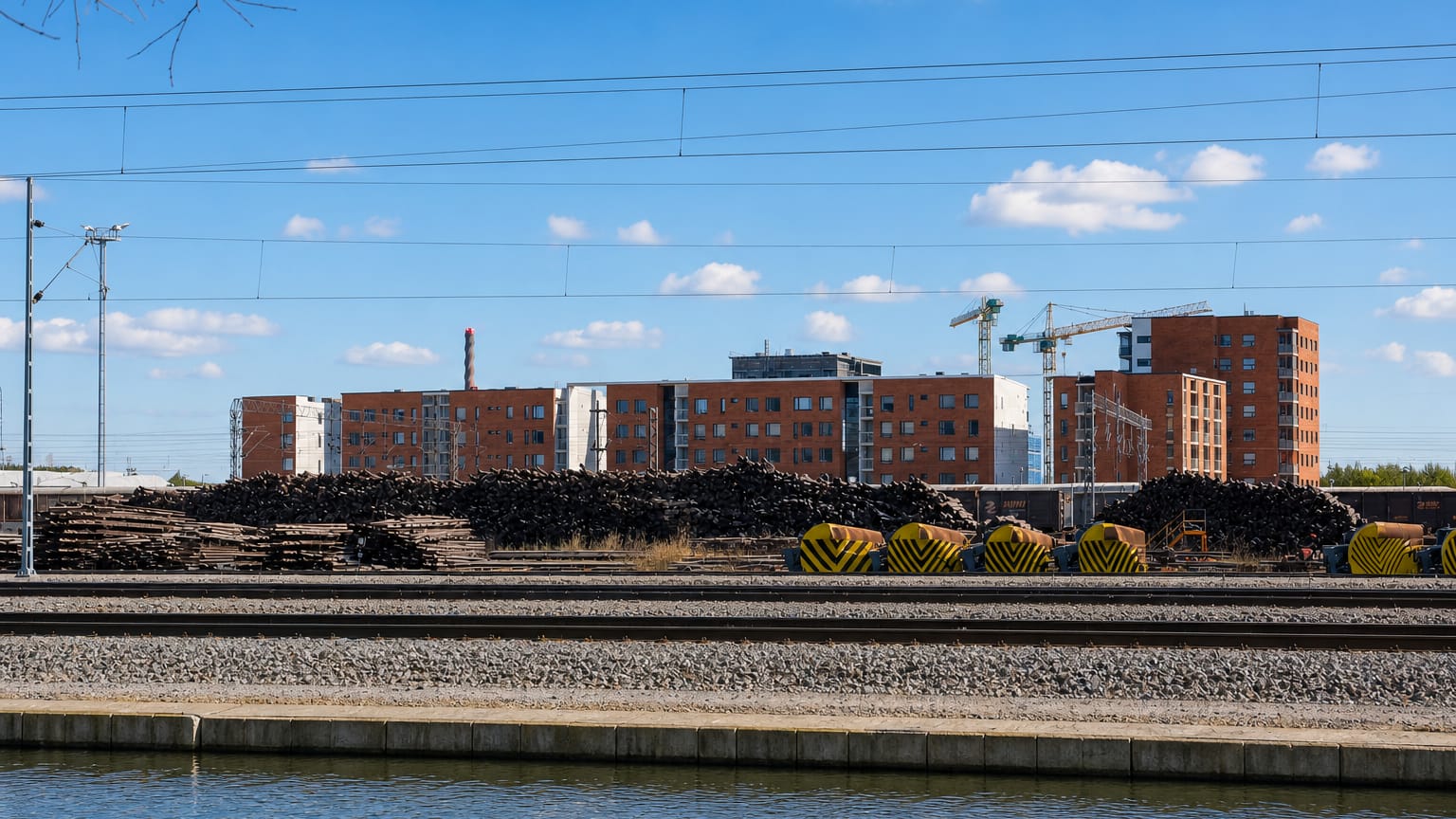 Red-brick apartment blocks behind electrified railway tracks, timber stacks and a canal in Limingantulli, Oulu, Finland.