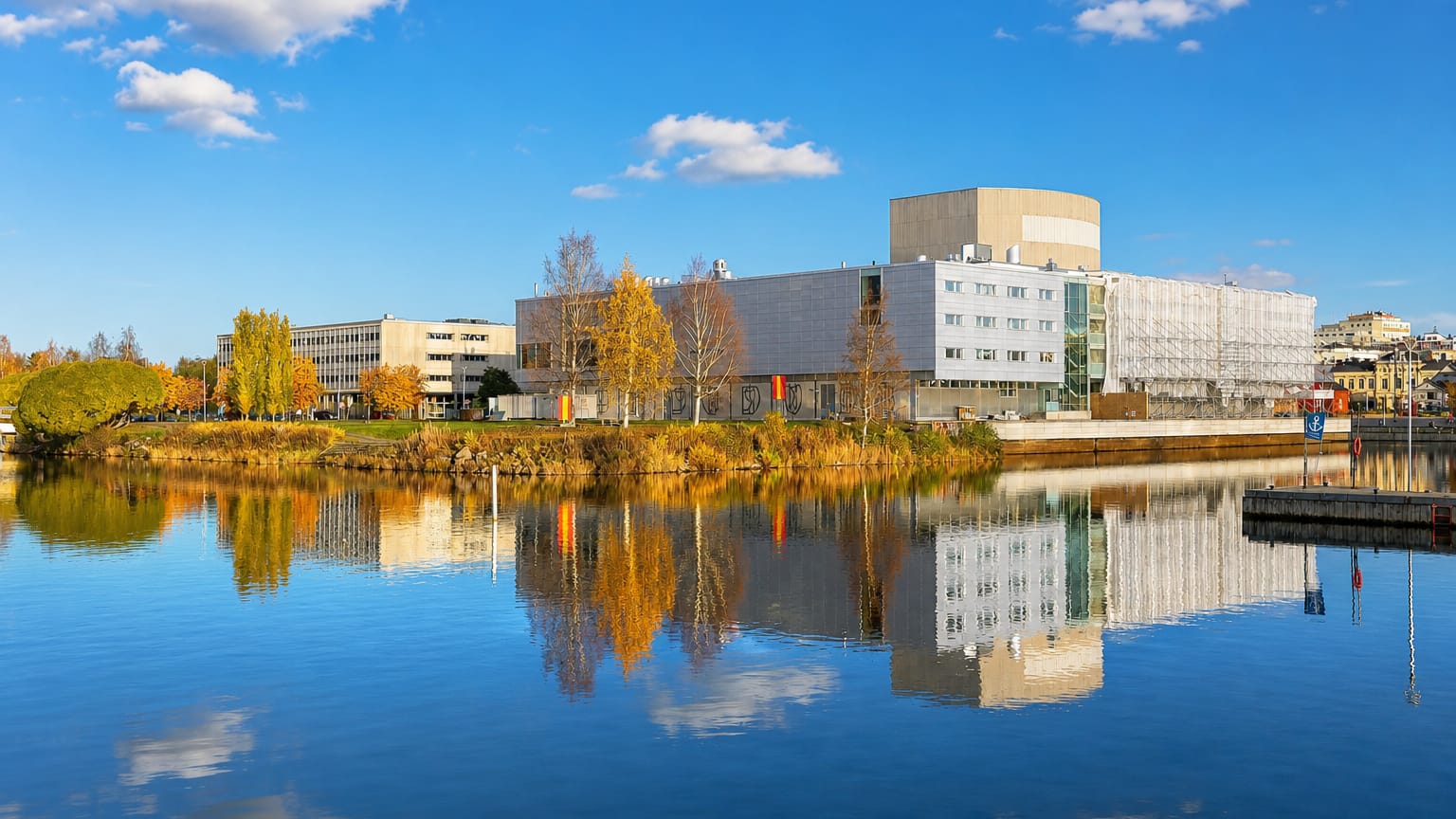 Pokkinen waterfront in Oulu, Finland, modern white buildings and autumn trees mirrored in calm river water under a blue sky