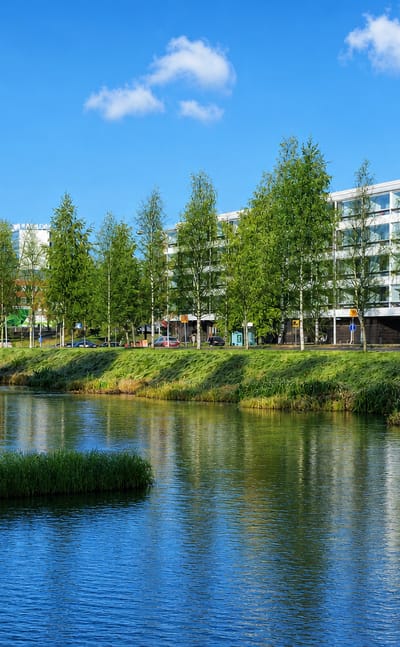 Calm waterway lined with birch trees reflecting modern mid-rise buildings under a blue sky in Raksila, Oulu, Finland.