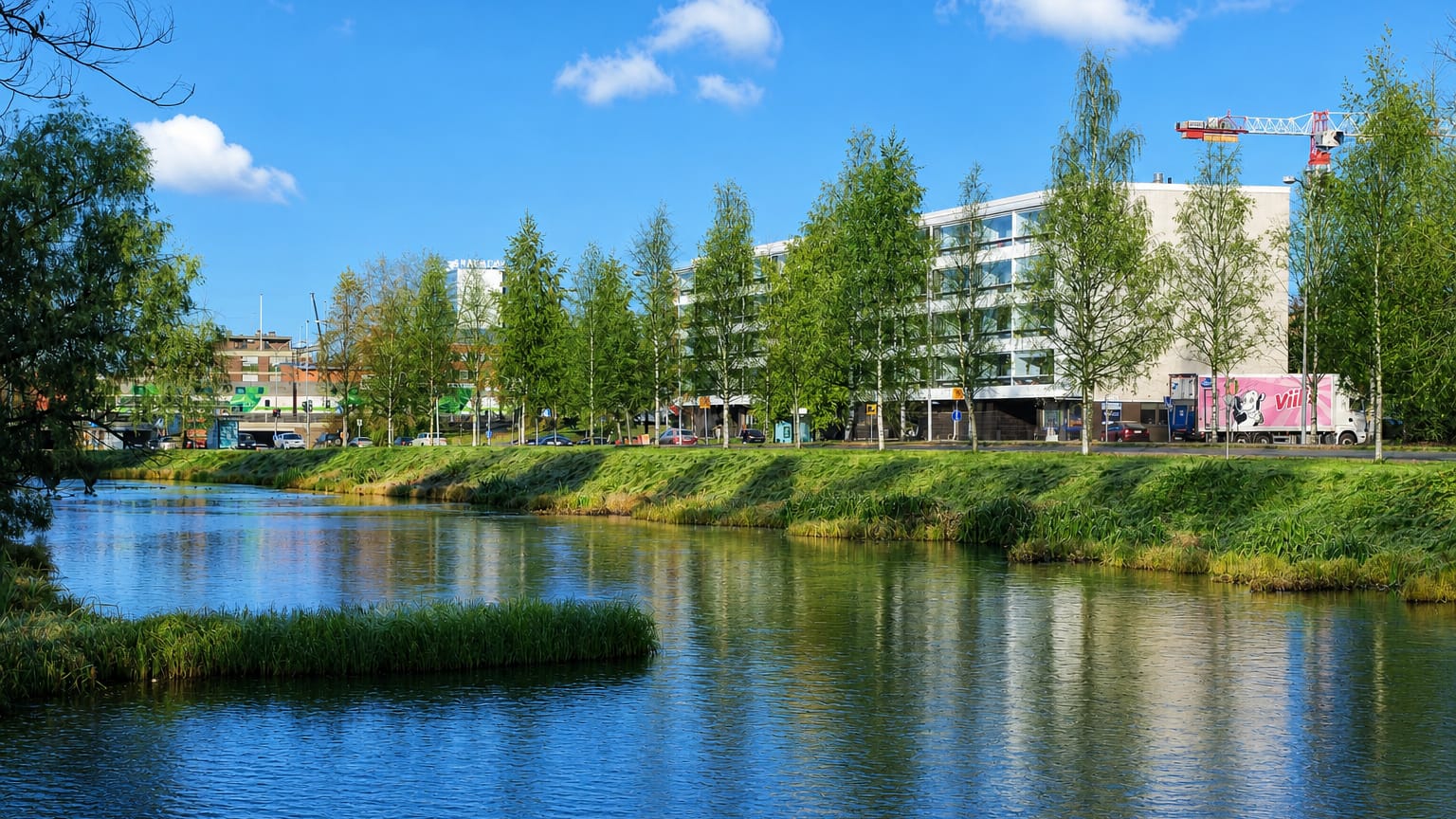 Calm waterway lined with birch trees reflecting modern mid-rise buildings under a blue sky in Raksila, Oulu, Finland.