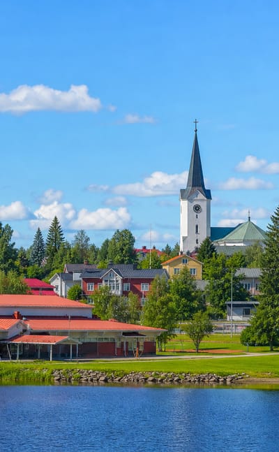 Pulkkila's white-spired church and red-roofed hall rise above a calm lakeshore under a bright summer sky.