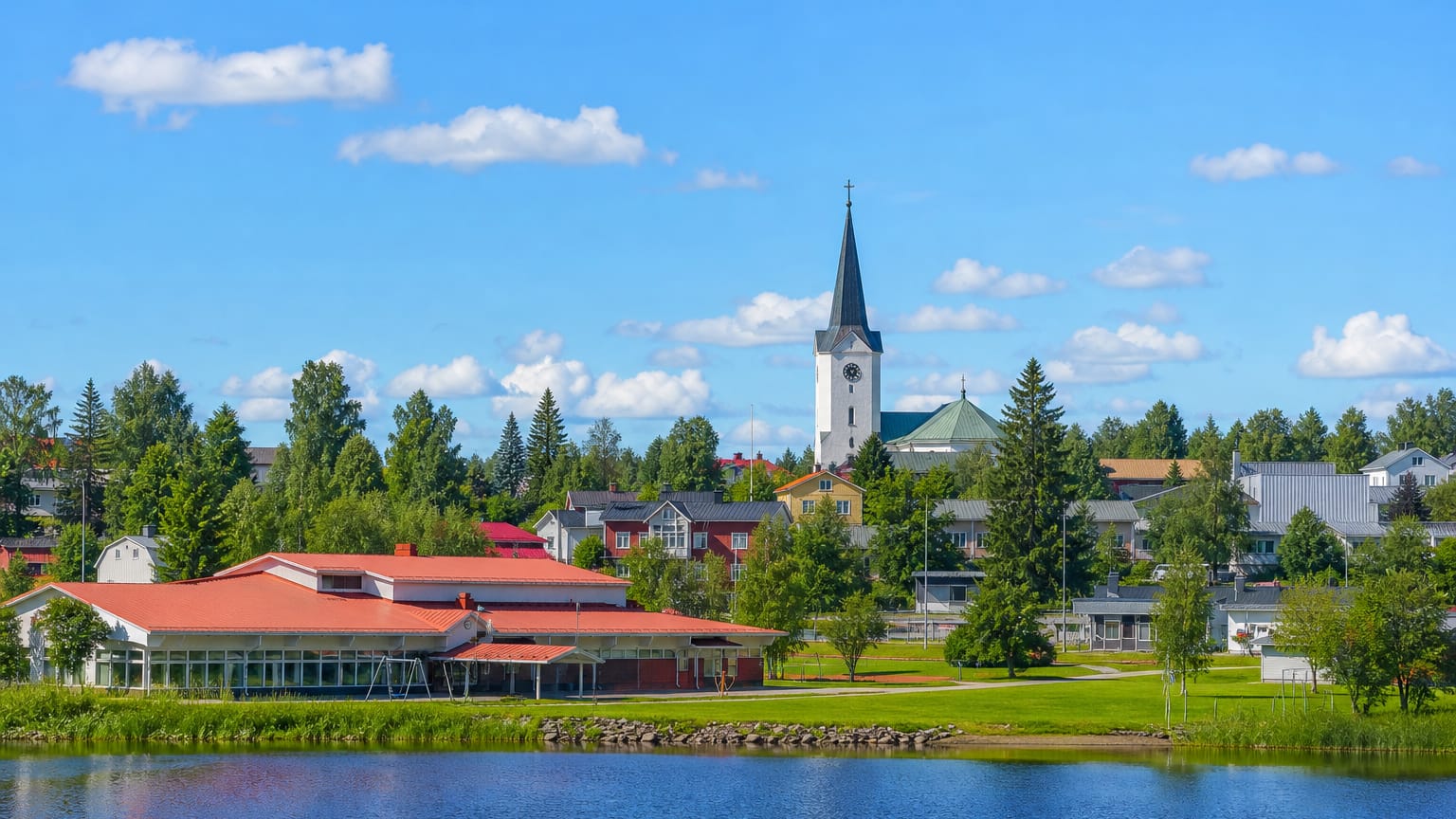 Pulkkila's white-spired church and red-roofed hall rise above a calm lakeshore under a bright summer sky.