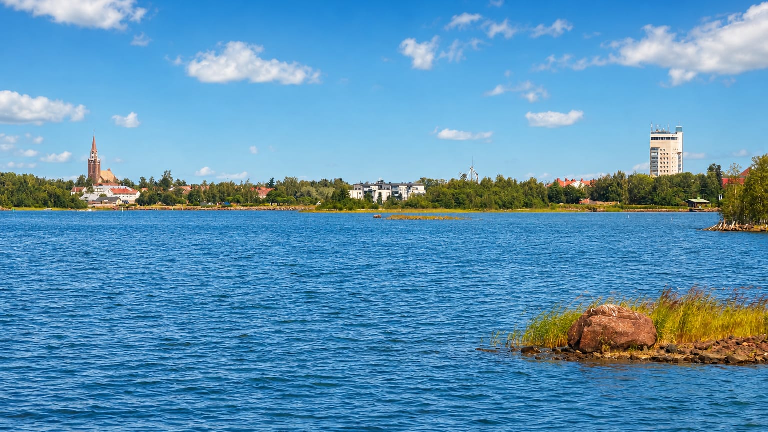 Raahe's low skyline across the blue Gulf of Bothnia, a church spire at left and a tall waterfront building at right under a summer sky.
