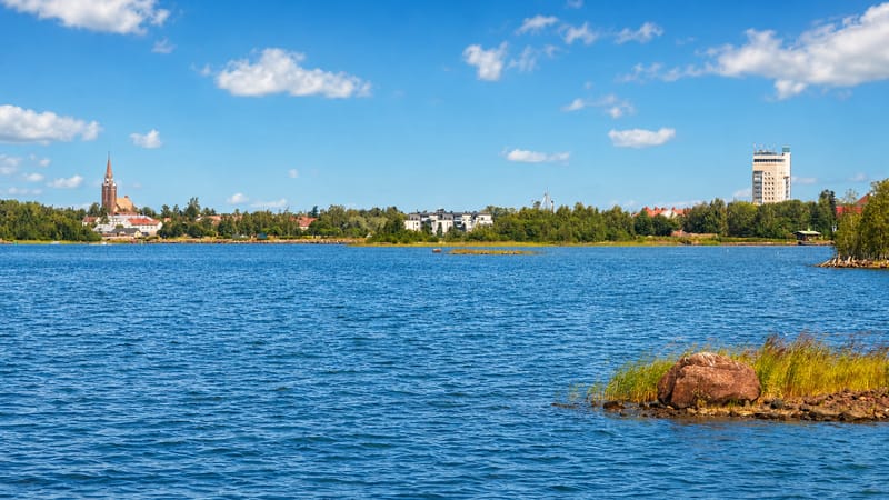 Raahe's low skyline across the blue Gulf of Bothnia, a church spire at left and a tall waterfront building at right under a summer sky.
