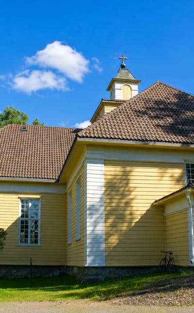 Joroinen's yellow wooden church with a small belfry and tiled roof, framed by birch trees on a sunny day.