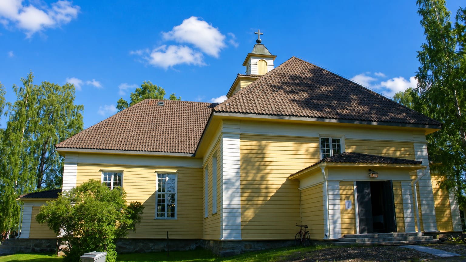 Joroinen's yellow wooden church with a small belfry and tiled roof, framed by birch trees on a sunny day.