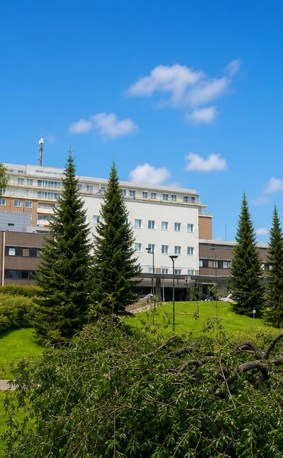 Large white hospital-style complex above a park with spruces and birches in Puijonlaakso, Kuopio.