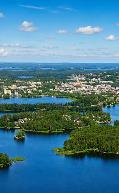 Aerial over the wooded islands of Rono on Lake Kallavesi, with Kuopio's centre and Puijo tower beyond.