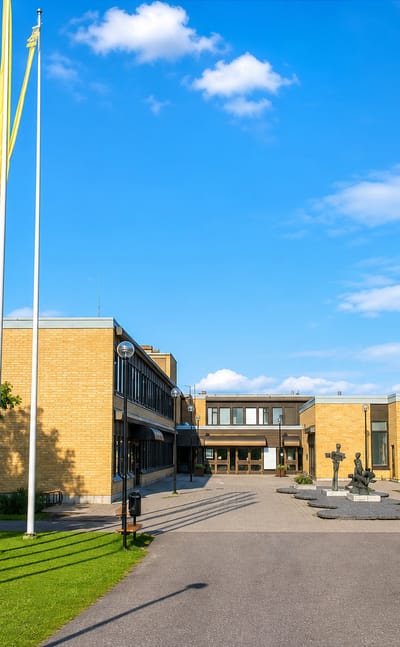 Modern yellow-brick civic building with flagpoles and a plaza sculpture in Leppävirta, Finland.