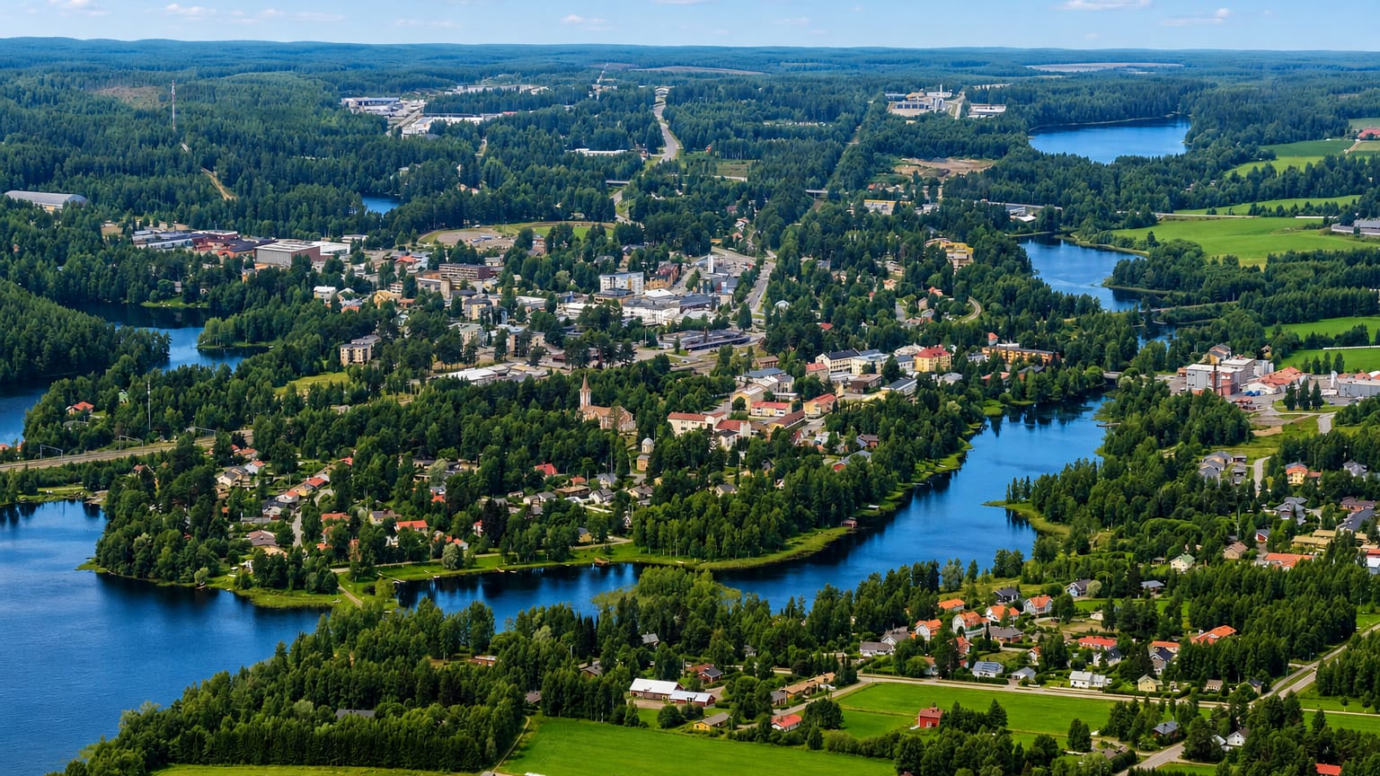 Aerial view of Suonenjoki, Finland, a small lakeland town ringed by blue lakes, green fields and forest under a bright summer sky.