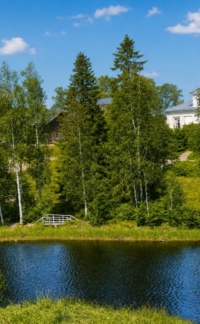 Summer lakeside in Vieremä, Finland, with white wooden buildings, a gazebo and footbridge among birch and spruce trees.