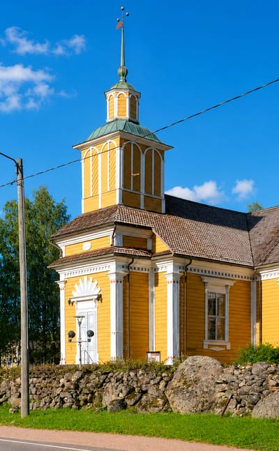 Yellow wooden church with a lantern tower behind a stone wall by a roadside in Honkajoki, Finland.
