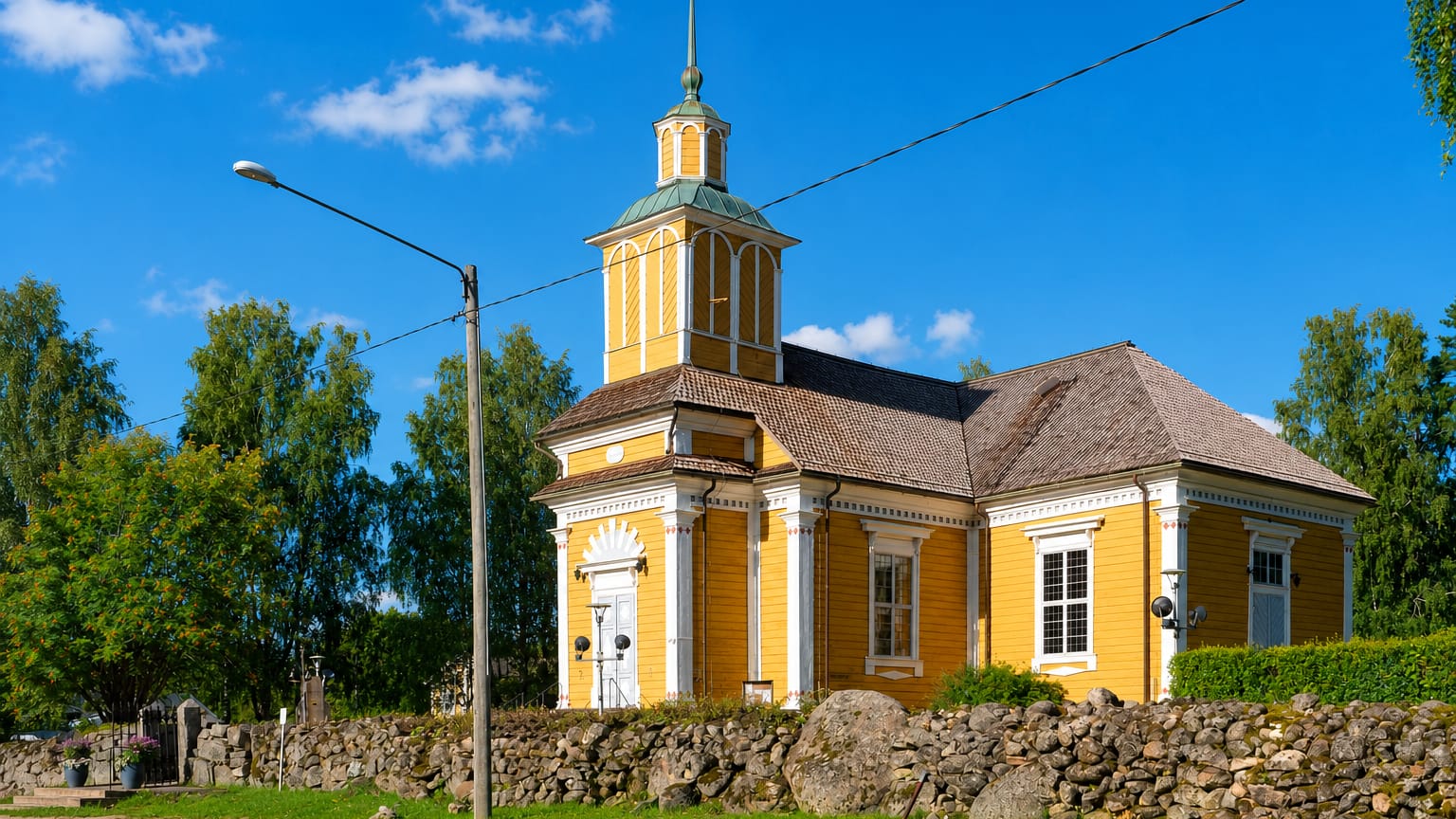 Yellow wooden church with a lantern tower behind a stone wall by a roadside in Honkajoki, Finland.