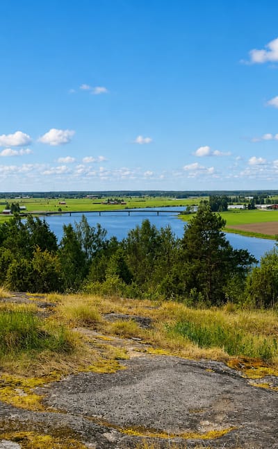 Rocky hilltop view over a river winding through flat farmland near Huittinen, Finland, under a blue summer sky