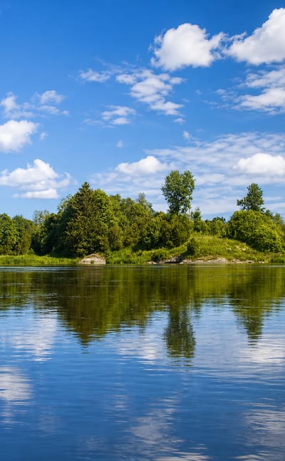 Calm river in Kokemäki, Finland, with a wooded islet and forested shore mirrored in still water under a blue summer sky.