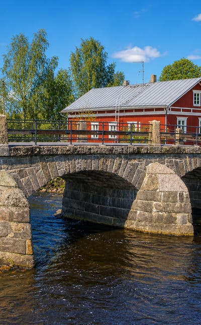 Three-arch stone bridge over the river in Pomarkku, Finland, with a red wooden building and birches under a blue summer sky.