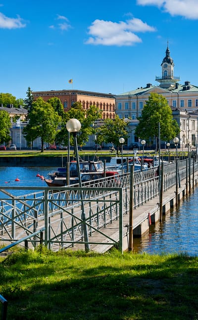 Riverside view in Pori with a pedestrian jetty, moored boats and historic buildings along the Kokemäenjoki under a blue summer sky.