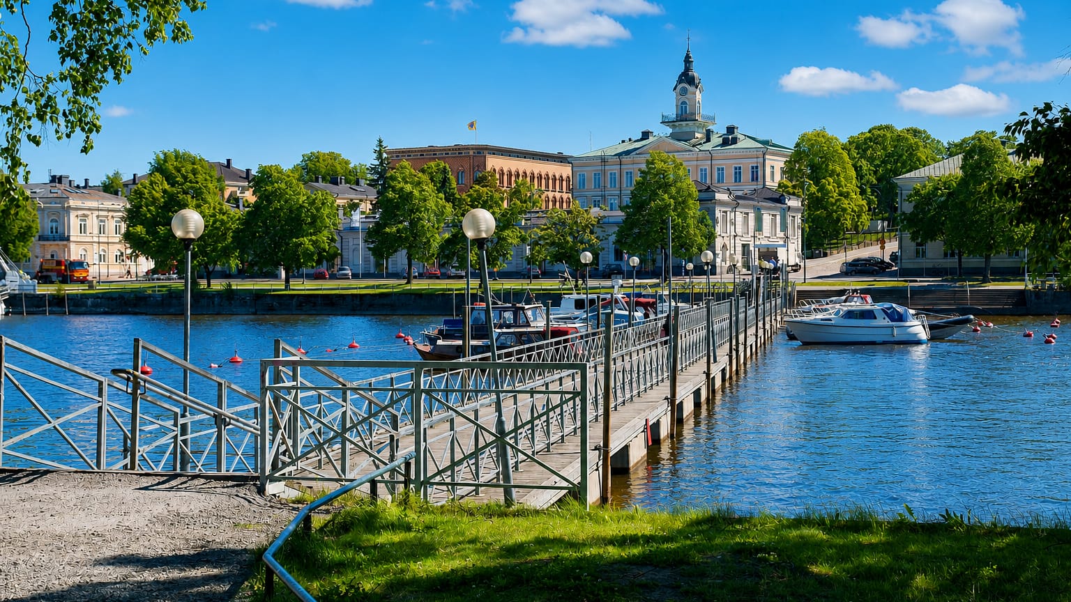 Riverside view in Pori with a pedestrian jetty, moored boats and historic buildings along the Kokemäenjoki under a blue summer sky.