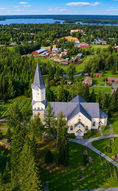 Aerial view of Siikainen's pale church and spire beside its cemetery, ringed by spruce forest and distant lakes, Satakunta, Finland.