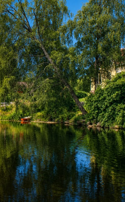 Calm river reflecting a yellow wooden house, red cabin and birch trees beside a road bridge in Luumäki, Finland.