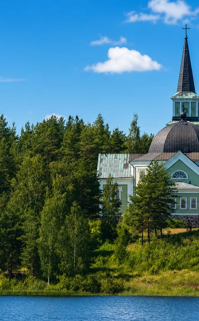 Pale wooden lakeside church with a tall steeple rising above dense pine forest in Ruokolahti, Finland.