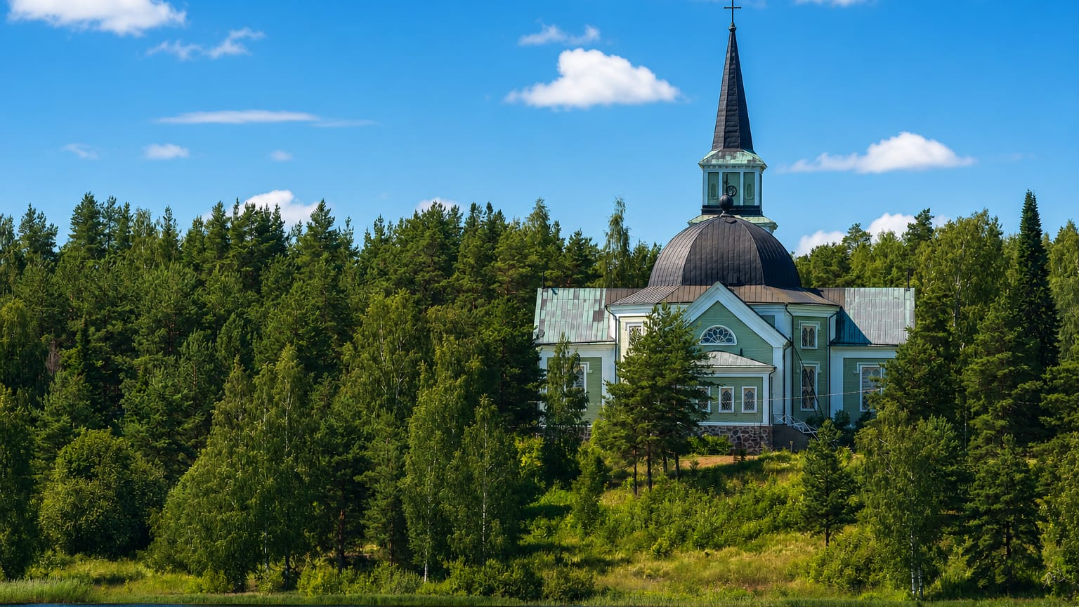 Pale wooden lakeside church with a tall steeple rising above dense pine forest in Ruokolahti, Finland.