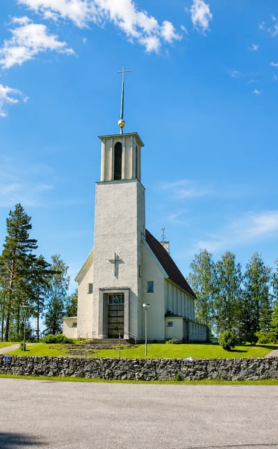 Simpele's white functionalist church with a tall bell tower and cross behind a stone wall and trees.