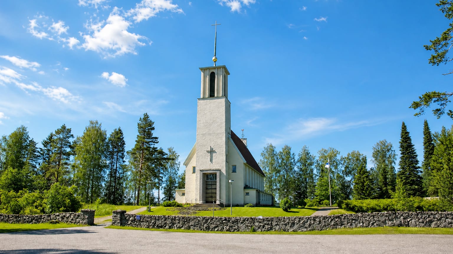Simpele's white functionalist church with a tall bell tower and cross behind a stone wall and trees.