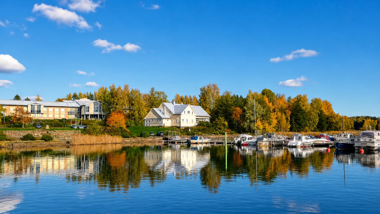 Lakeside marina at Taipalsaari, Finland, with small boats moored on calm Lake Saimaa beneath autumn birch trees.