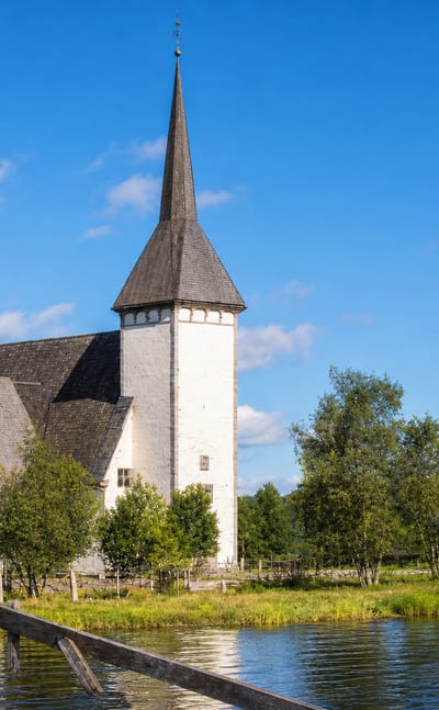 White stone church with a tall dark spire and separate bell tower beside a lake among birches in Alahärmä, Finland.