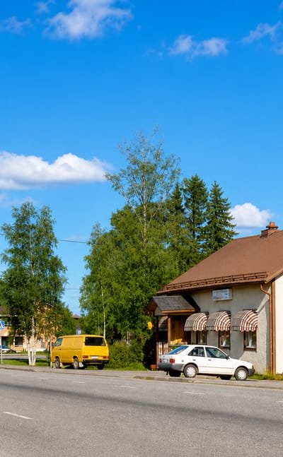 Roadside house with striped awnings, parked cars and birch trees along a curving road in Karijoki, Finland.