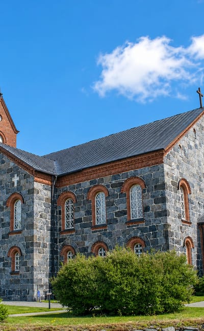 Juva's grey fieldstone church with red-brick trim and a slender spire under a bright blue sky.