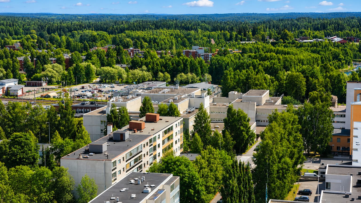 Aerial view over Maunuksela in Mikkeli: flat-roofed apartment blocks among birch woods and forested ridges.