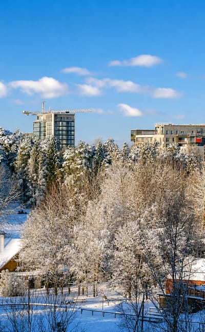 Snow-covered forest and a wooden house below modern apartment towers and construction cranes in Espoonlahti, Espoo, Finland