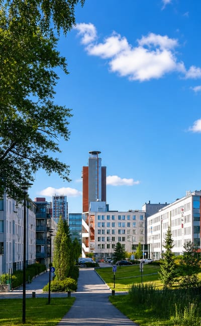 Modern apartment and office blocks and a slim tower beyond a birch-lined park path in Leppävaara, Espoo, Finland