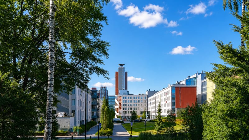 Modern apartment and office blocks and a slim tower beyond a birch-lined park path in Leppävaara, Espoo, Finland
