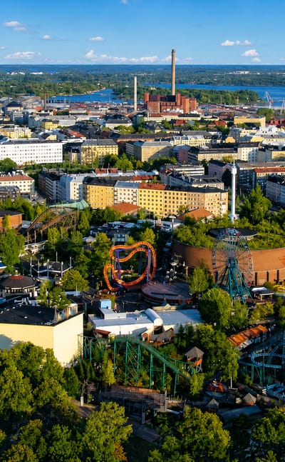 Aerial view of Alppiharju, Helsinki, with Linnanmäki amusement park's roller coasters amid greenery and the city skyline beyond.